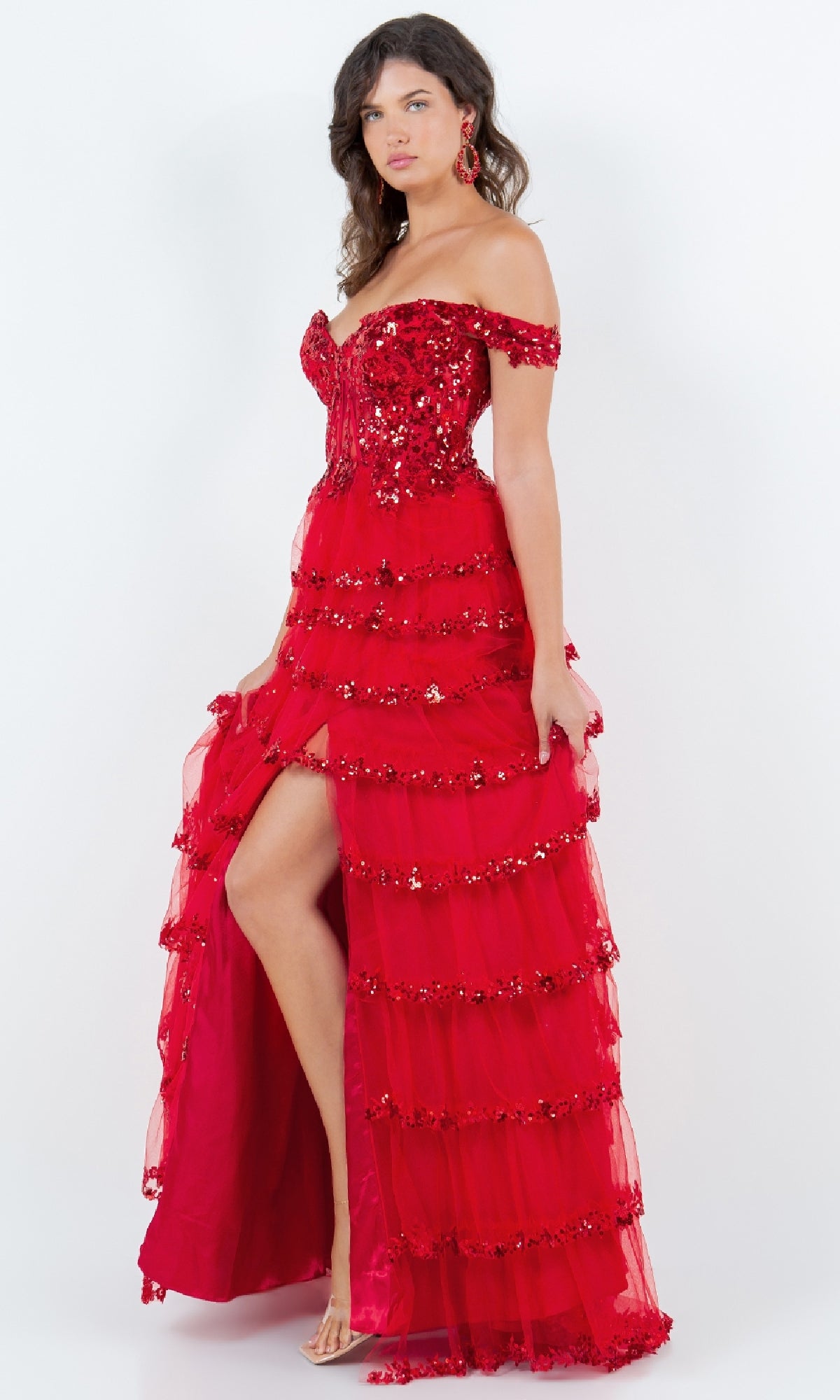A person poses against a plain white background in the Cinderella Couture 8228J Ruffle Prom Ball Gown—a red, off-the-shoulder dress with sequin embroidery, tiered ruffles, and a dramatic thigh-high slit.